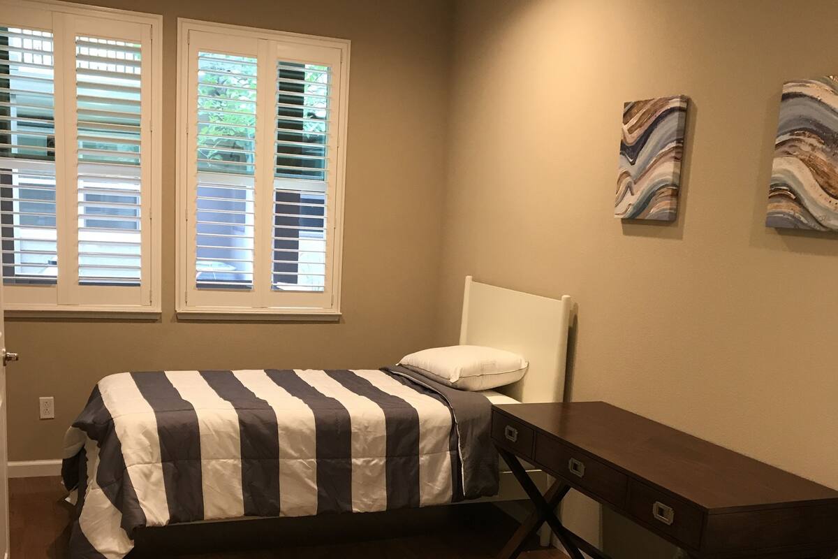 A bedroom with a bed, a desk, and two windows with blinds.