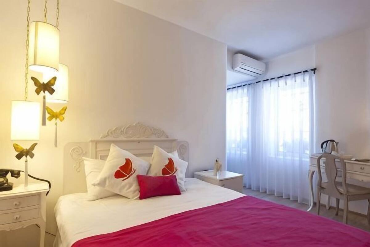 A hotel room with a bed, a desk, a chair, a nightstand, and an air conditioning unit.