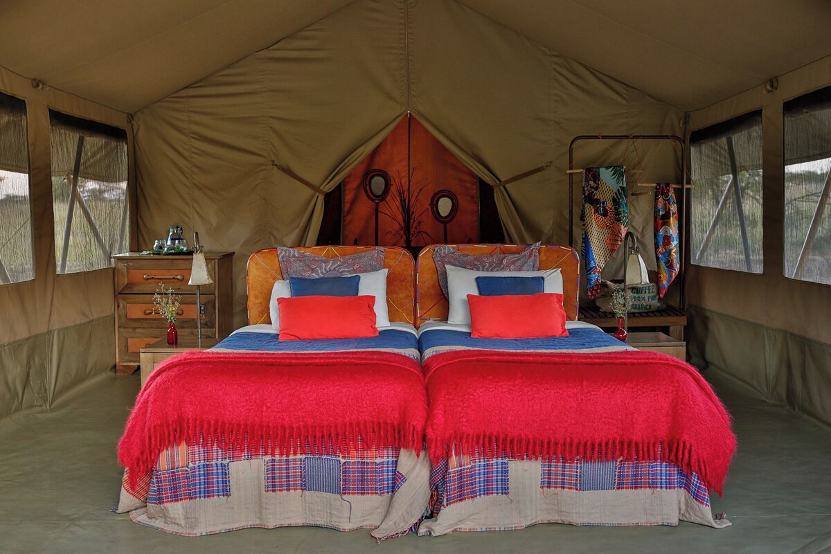 Uma tenda com duas camas, cada uma com roupa de cama vermelha e azul, e um aparador de madeira com um espelho.