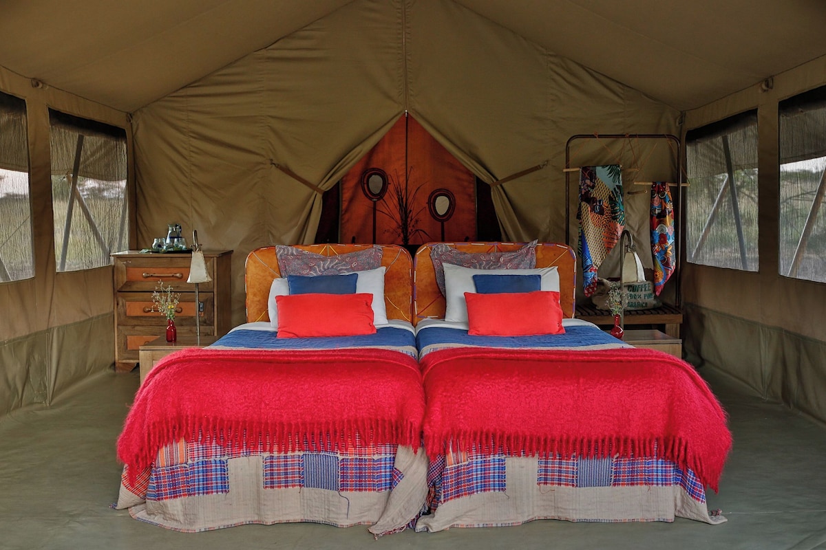 A tent with two beds, each with red and blue bedding, and a wooden dresser with a mirror.