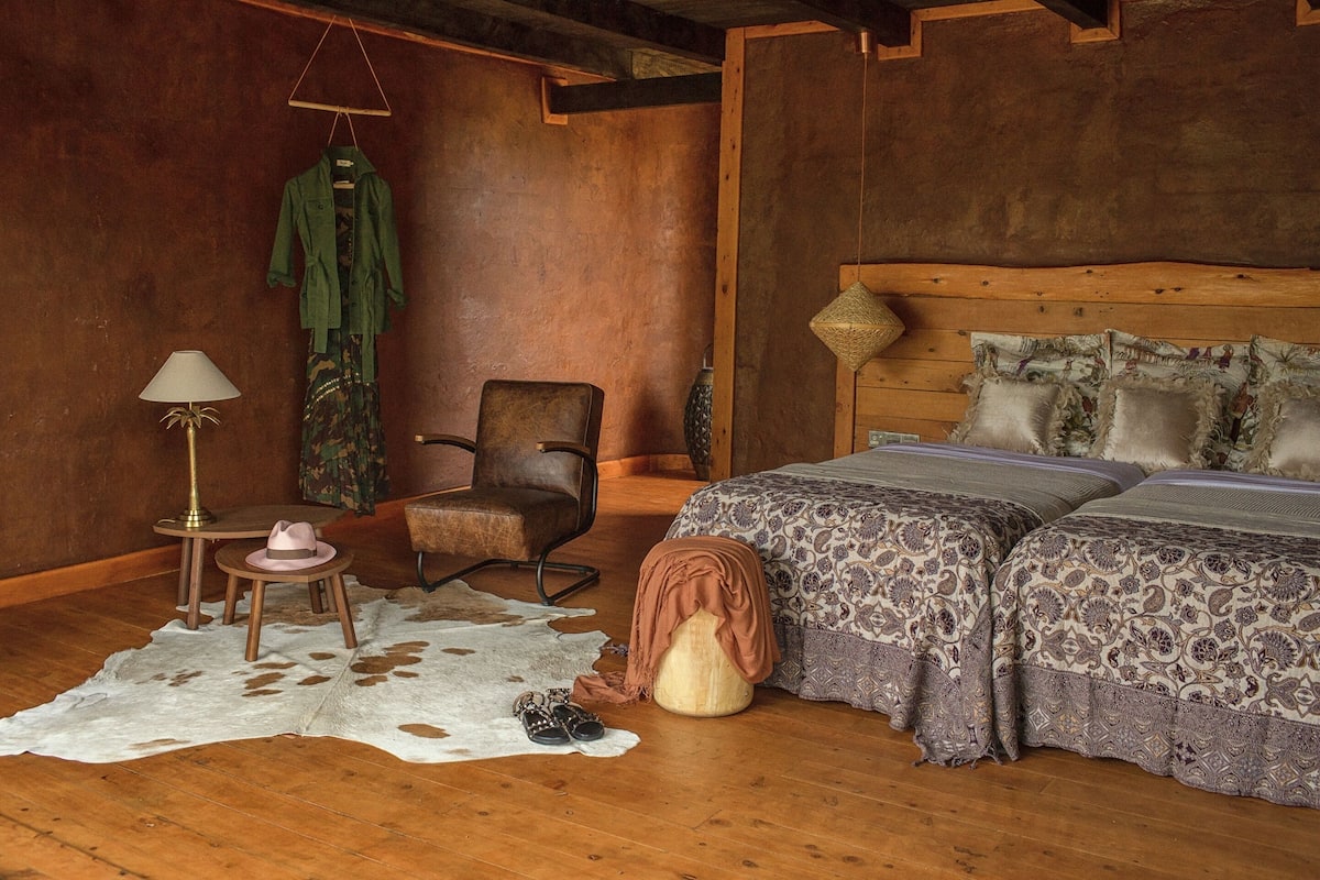 A rustic bedroom with a wooden headboard, a large bed with patterned bedding, a chair, a lamp, and a coat hanging on the wall.