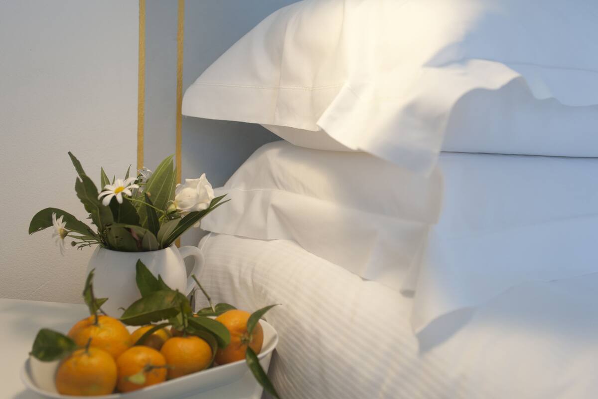 A white bed with pillows, a white vase with flowers and oranges on a bedside table.