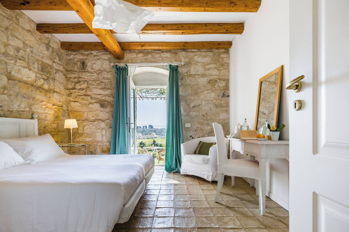 A bedroom with stone walls, a wooden ceiling, a bed, a desk, and a chair.
