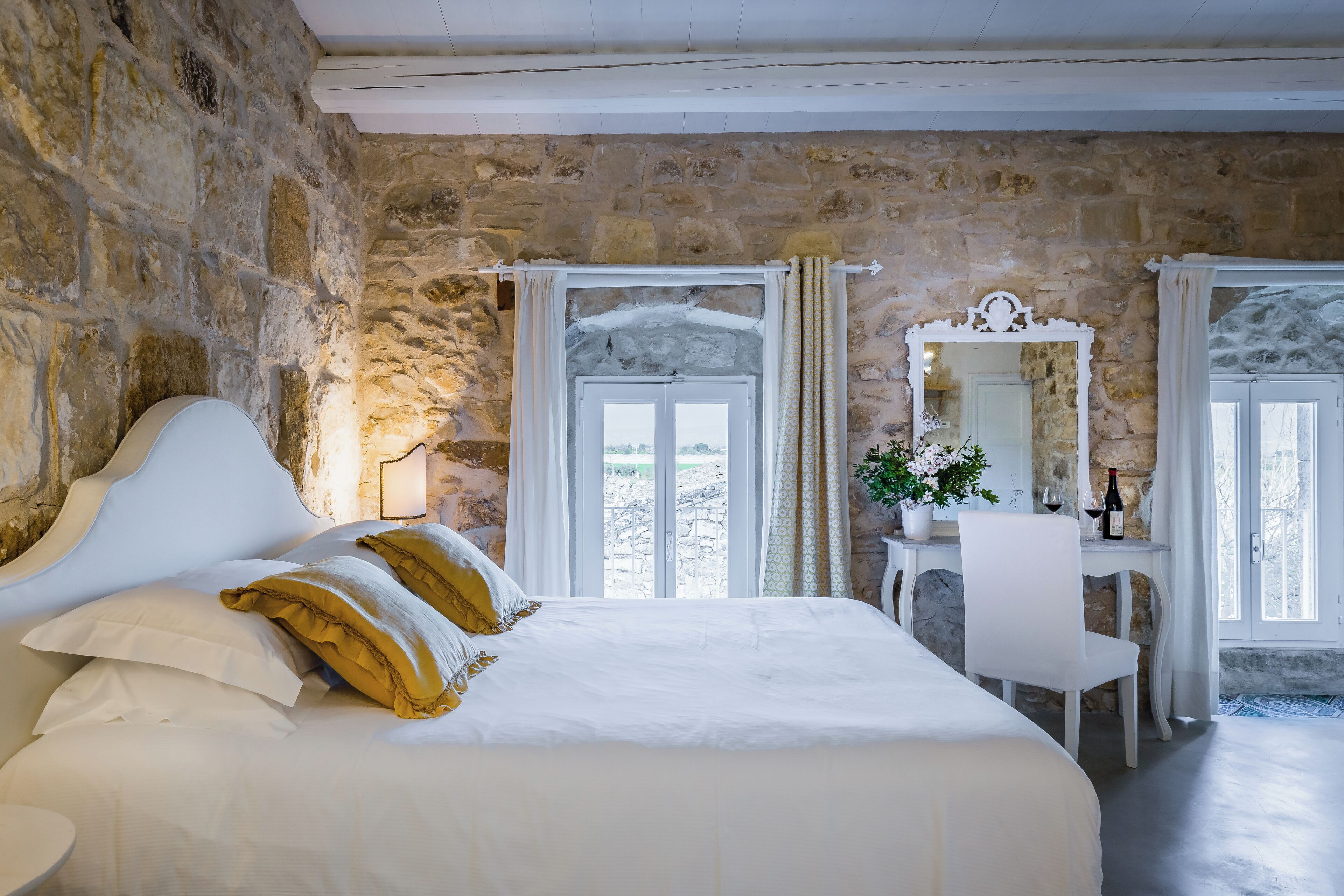 A bedroom with a stone wall, a bed with white and yellow pillows, a desk with a chair, and a mirror.