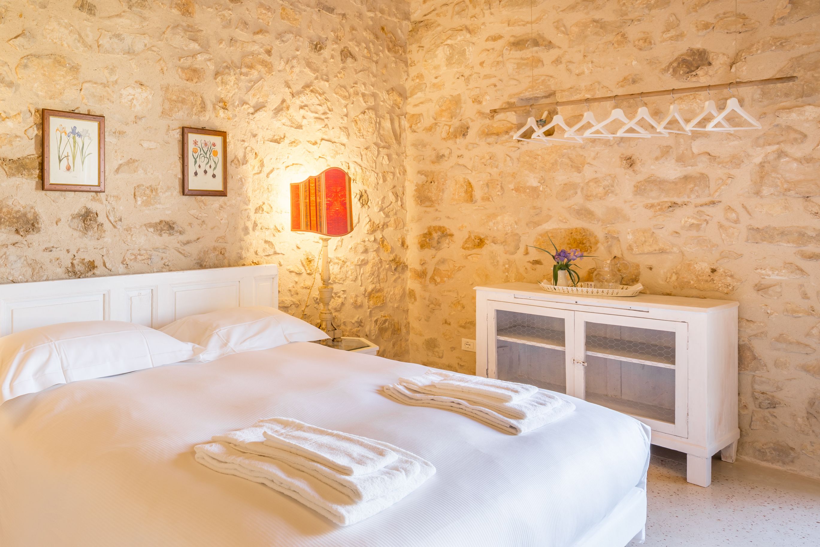 A bedroom with a stone wall, a bed with white linens, a nightstand, a lamp, and a cabinet with a vase of flowers.