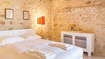 A bedroom with a stone wall, a bed with white linens, a nightstand, a lamp, and a cabinet with a vase of flowers.
