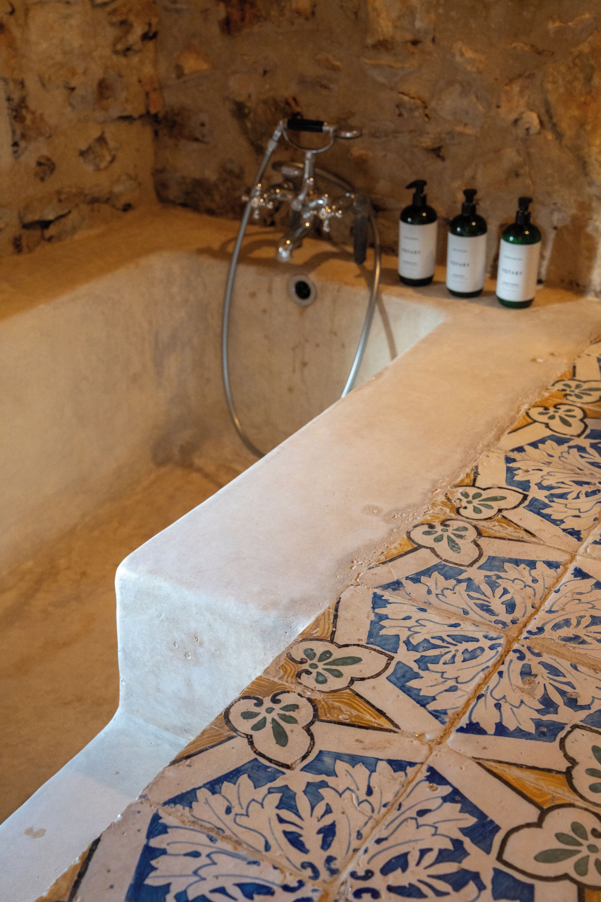 A bathtub with a tiled edge and a showerhead, three bottles of shampoo, and a stone wall in the background.