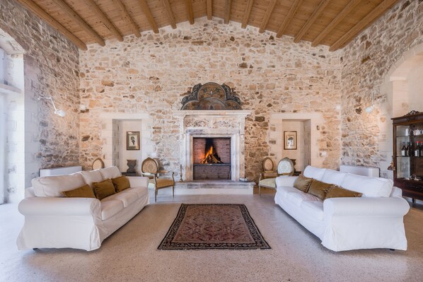 Una sala de estar con paredes de piedra, una chimenea, dos sofás blancos y un tapete estampado.
