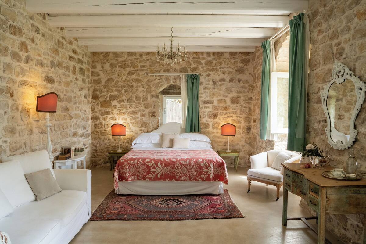 A bedroom with a stone wall, a bed with a red and white bedspread, a white sofa, a wooden desk, and a chair.
