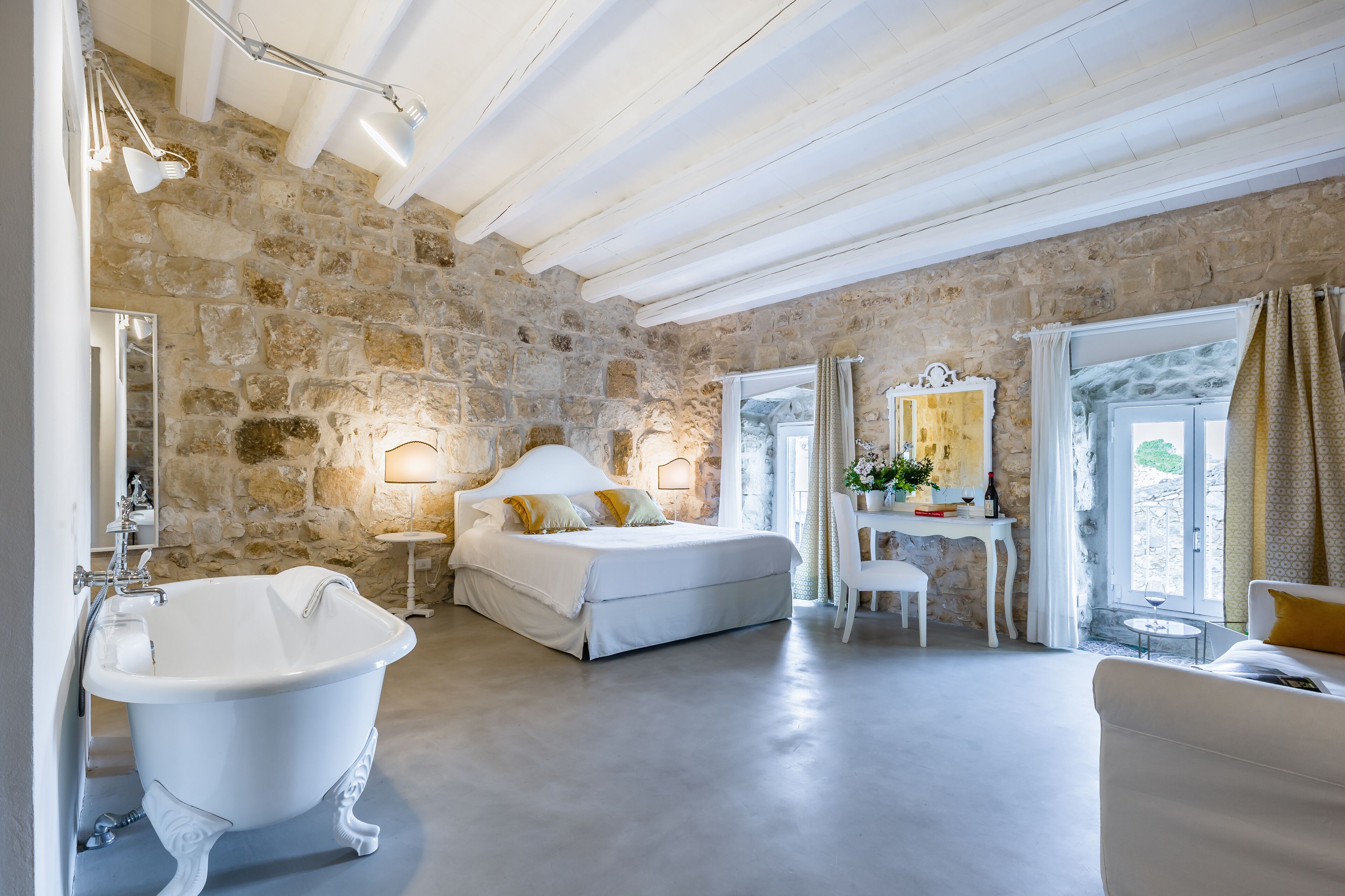 A spacious bedroom with a stone wall, a large bed, a white vanity, and a bathtub.
