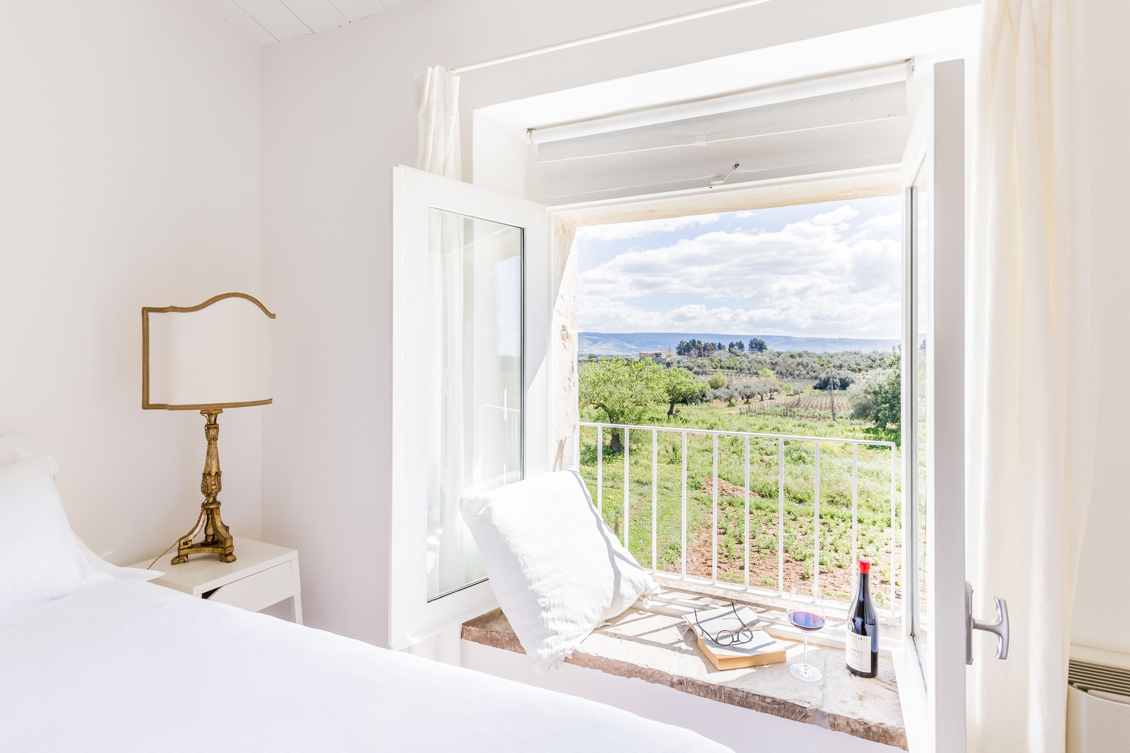 A bedroom with a large window, a bedside table with a lamp, a white pillow, and a bottle of wine.