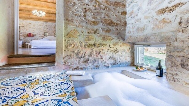Una habitación con paredes de piedra, una bañera grande, suelo estampado y ventana con vistas al agua.