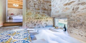 Una habitación con paredes de piedra, una bañera grande, suelo estampado y ventana con vistas al agua.