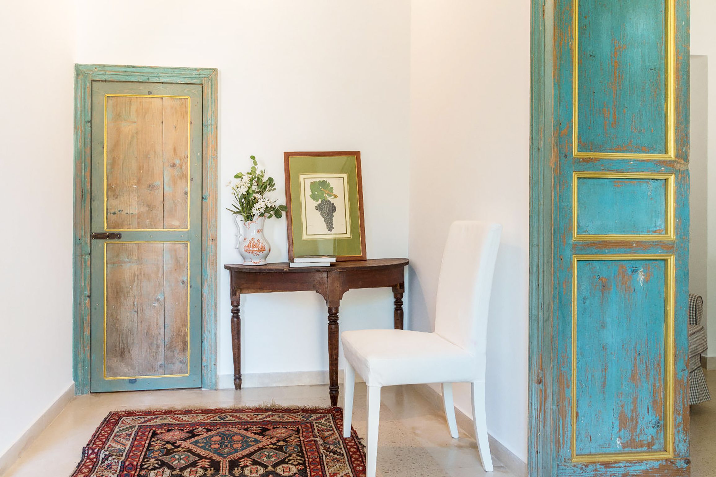 Una habitación con una silla blanca, una mesa de madera con un florero y un cuadro enmarco, y dos puertas con un acabado rústico.