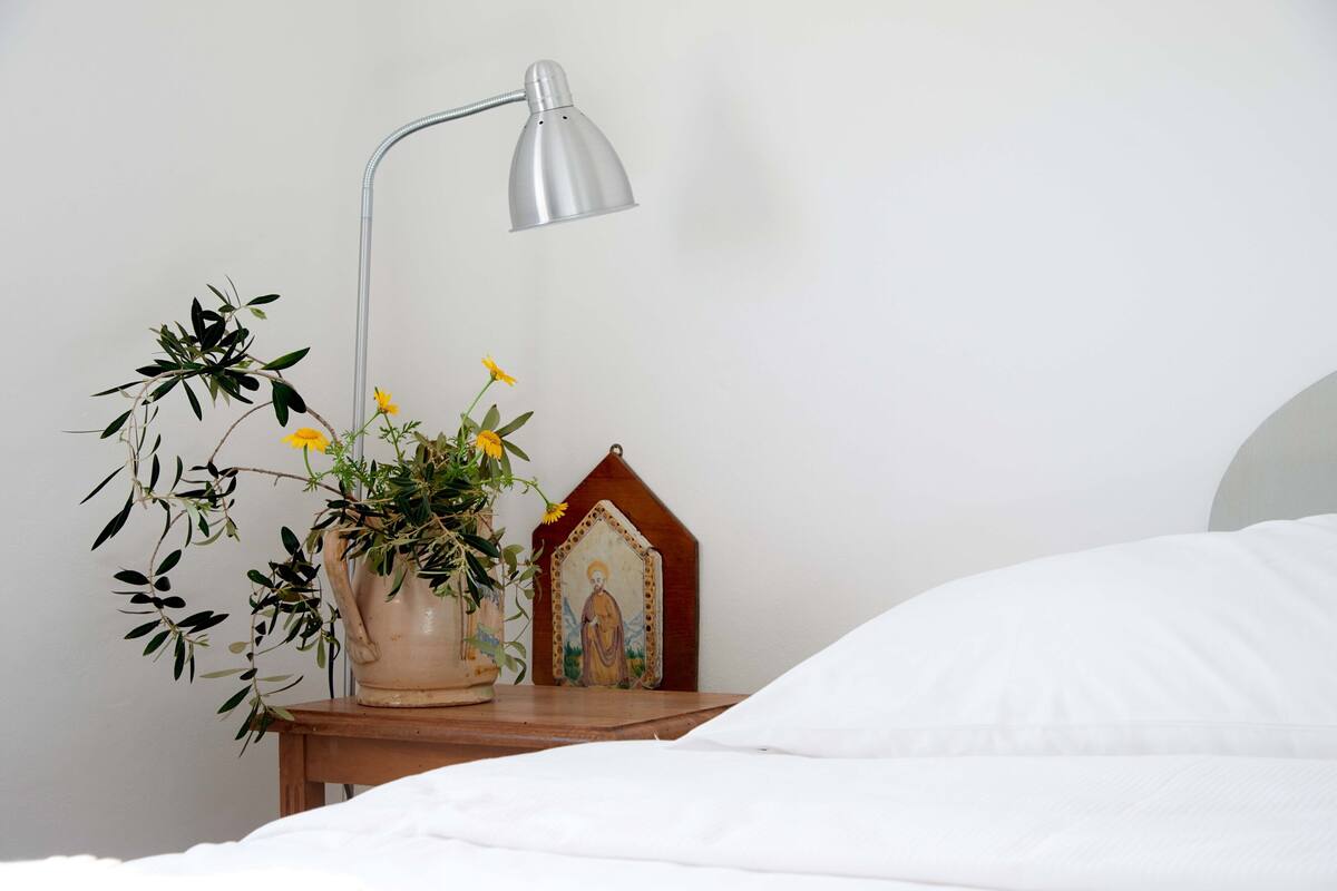 A neatly made bed with white sheets, a bedside table with a lamp, a potted plant, and a religious icon.