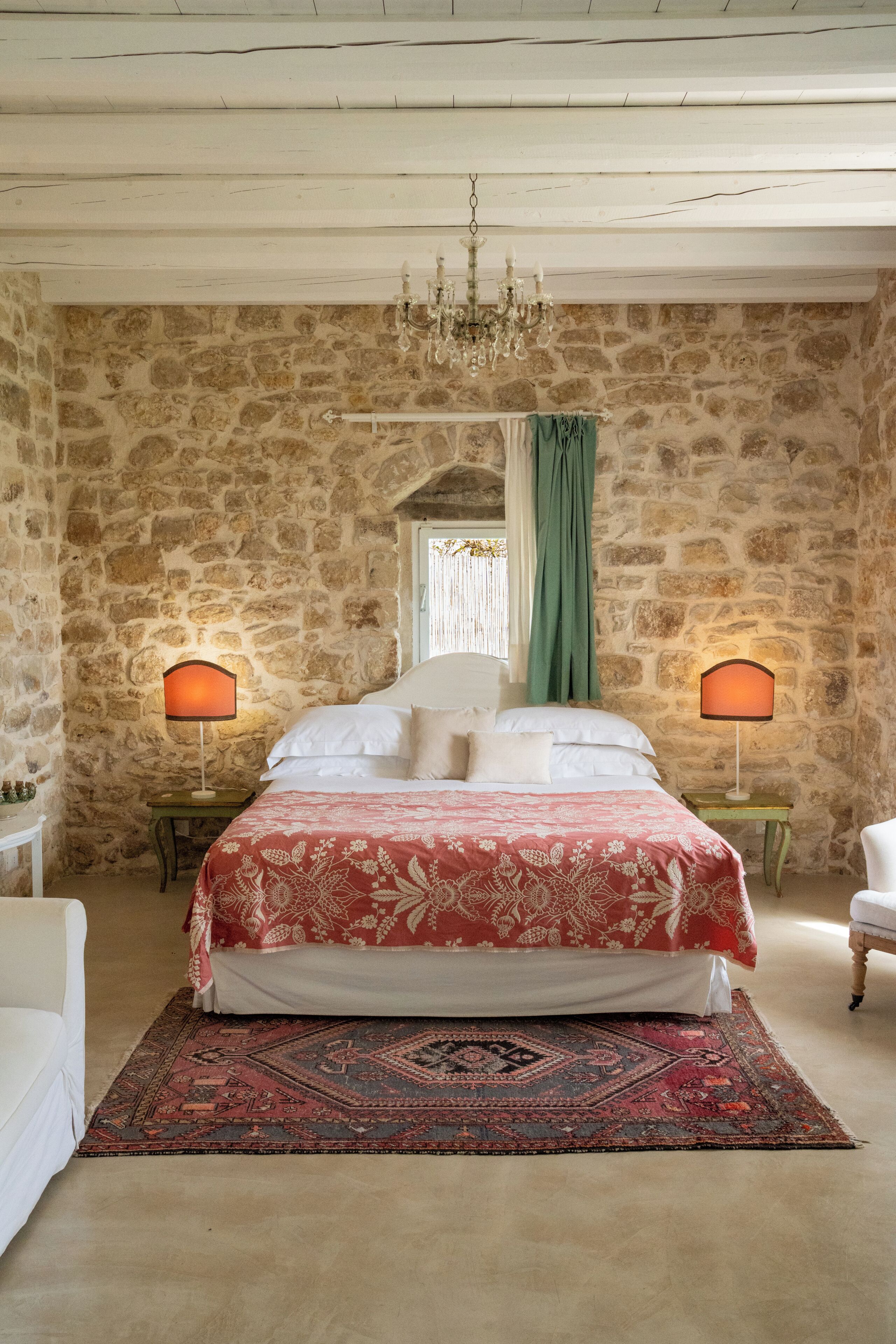 Une chambre avec un mur en pierre, un lustre, un lit recouvert d’une courtepointe rouge et blanche, deux lampes de chevet, un rideau vert et un tapis à motifs.