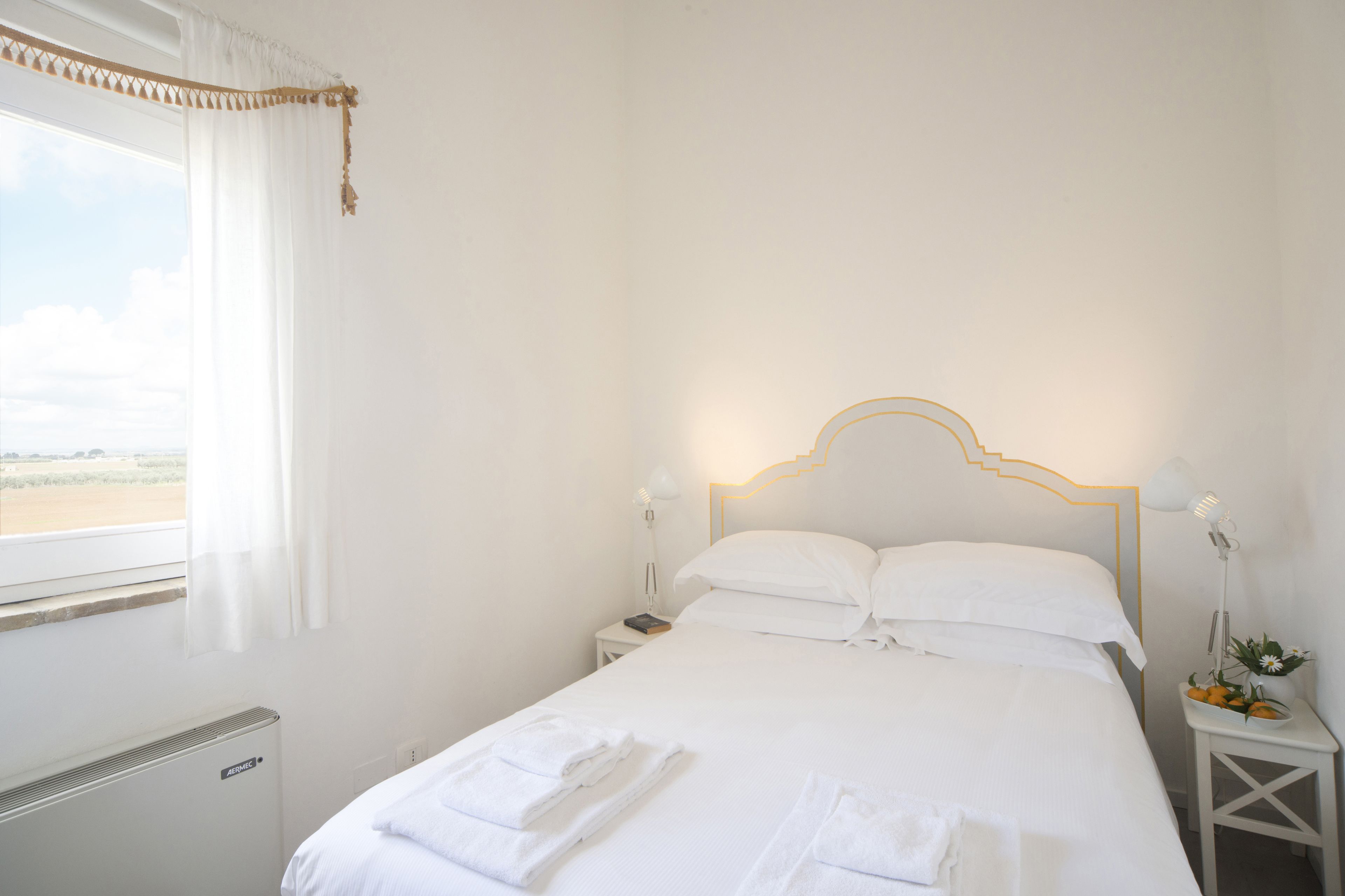 Una cama bien hecha con sábanas y toallas blancas, una mesita de noche con lámpara y vistas al exterior a través de una ventana con cortinas.