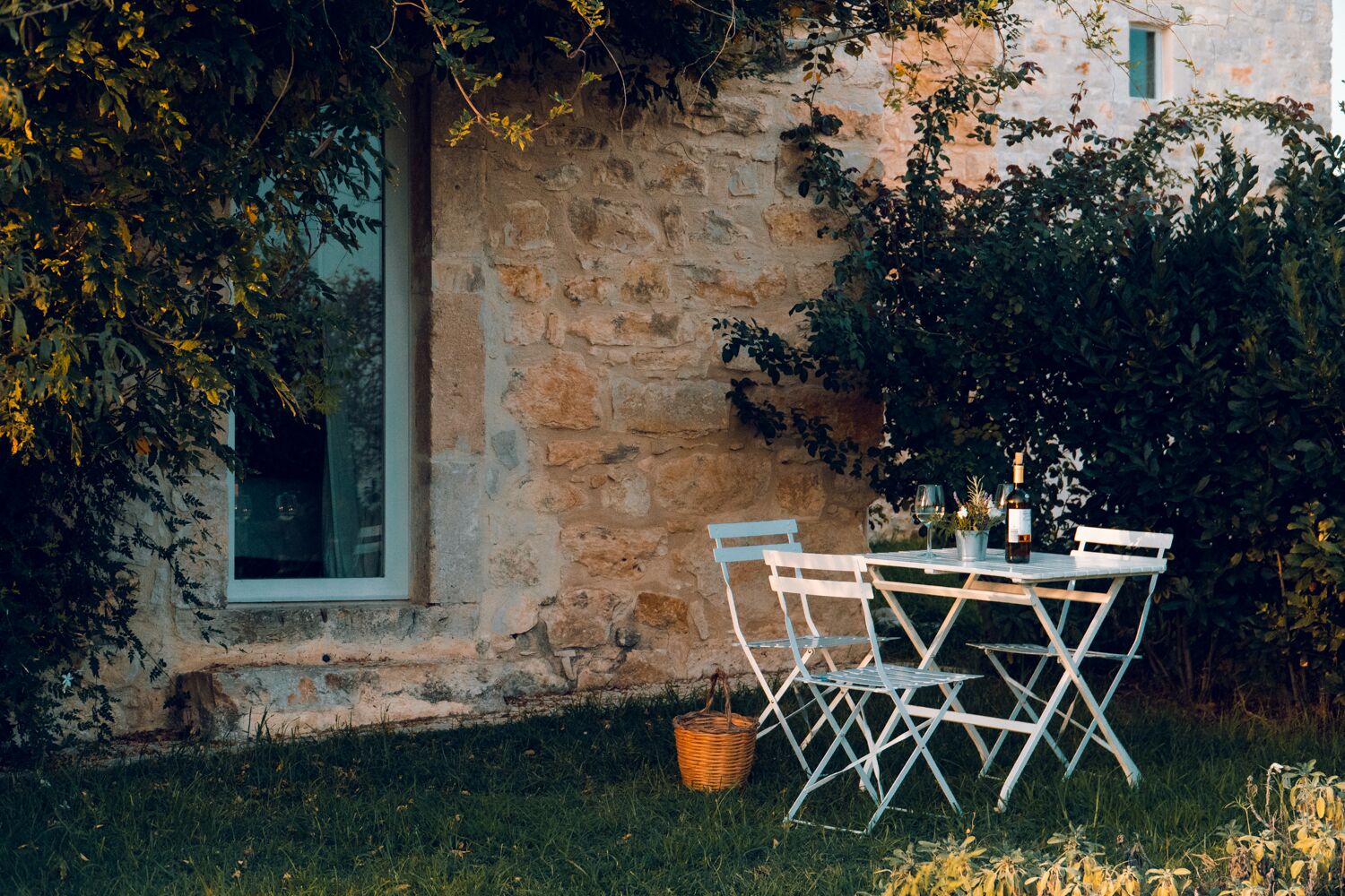 An outdoor dining area with a stone wall, a window, and a table set with chairs and a basket.
