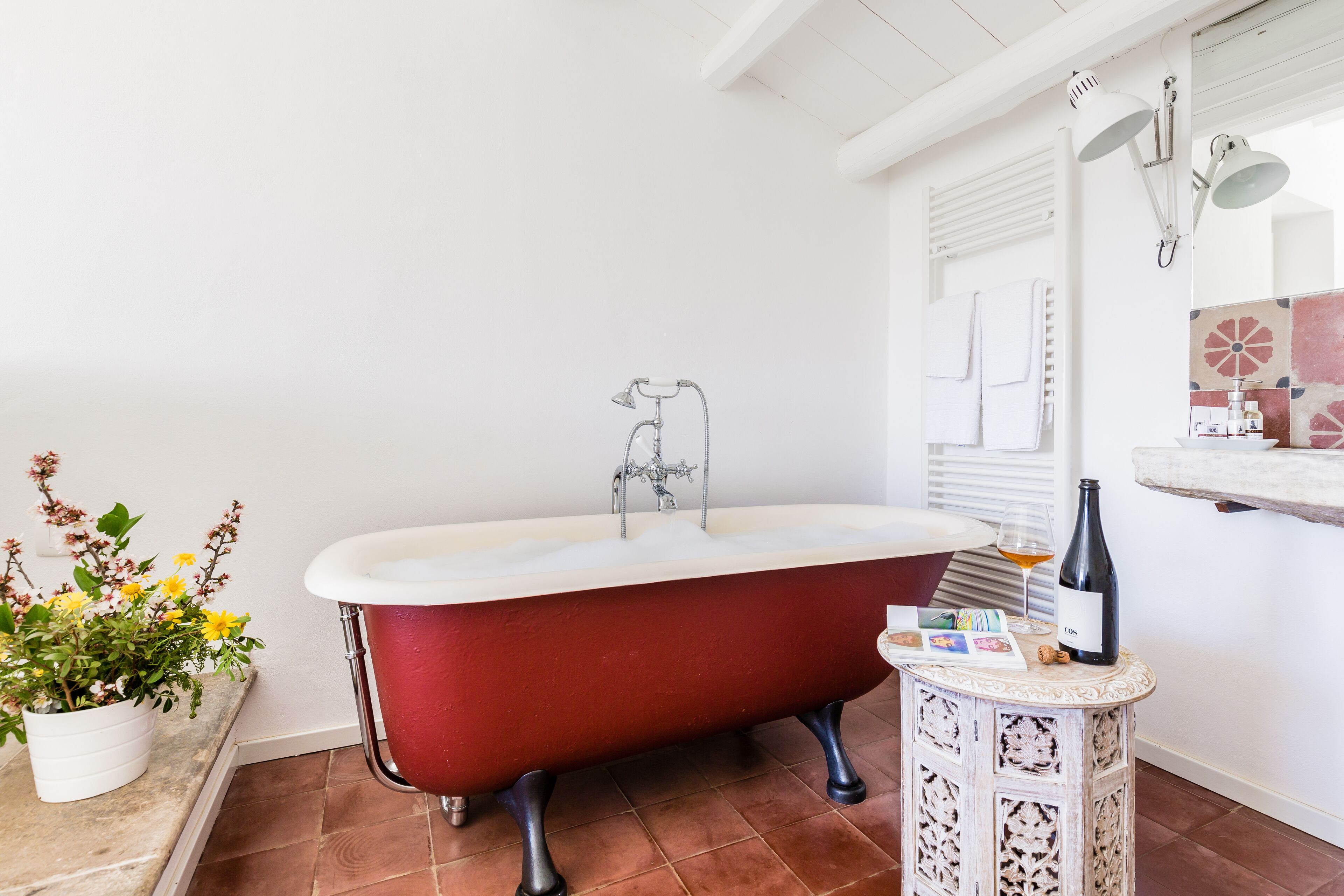 Un baño con una bañera roja tipo pata de león, un lavabo blanco, una pequeña mesa con una botella y vasos, y una planta en maceta.