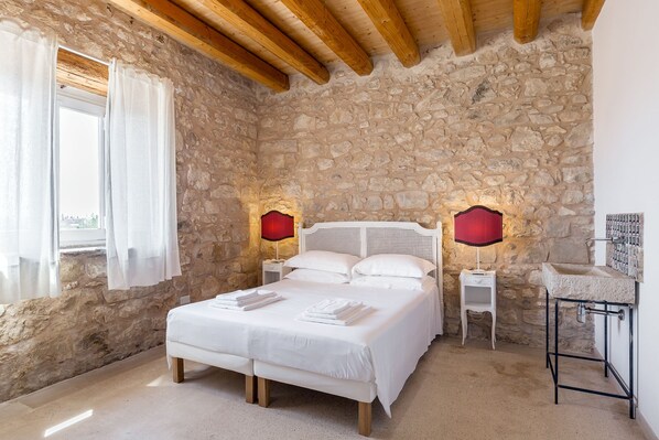 A bedroom with a stone wall, wooden ceiling beams, a white bed, two bedside tables with lamps, and a small white cabinet.