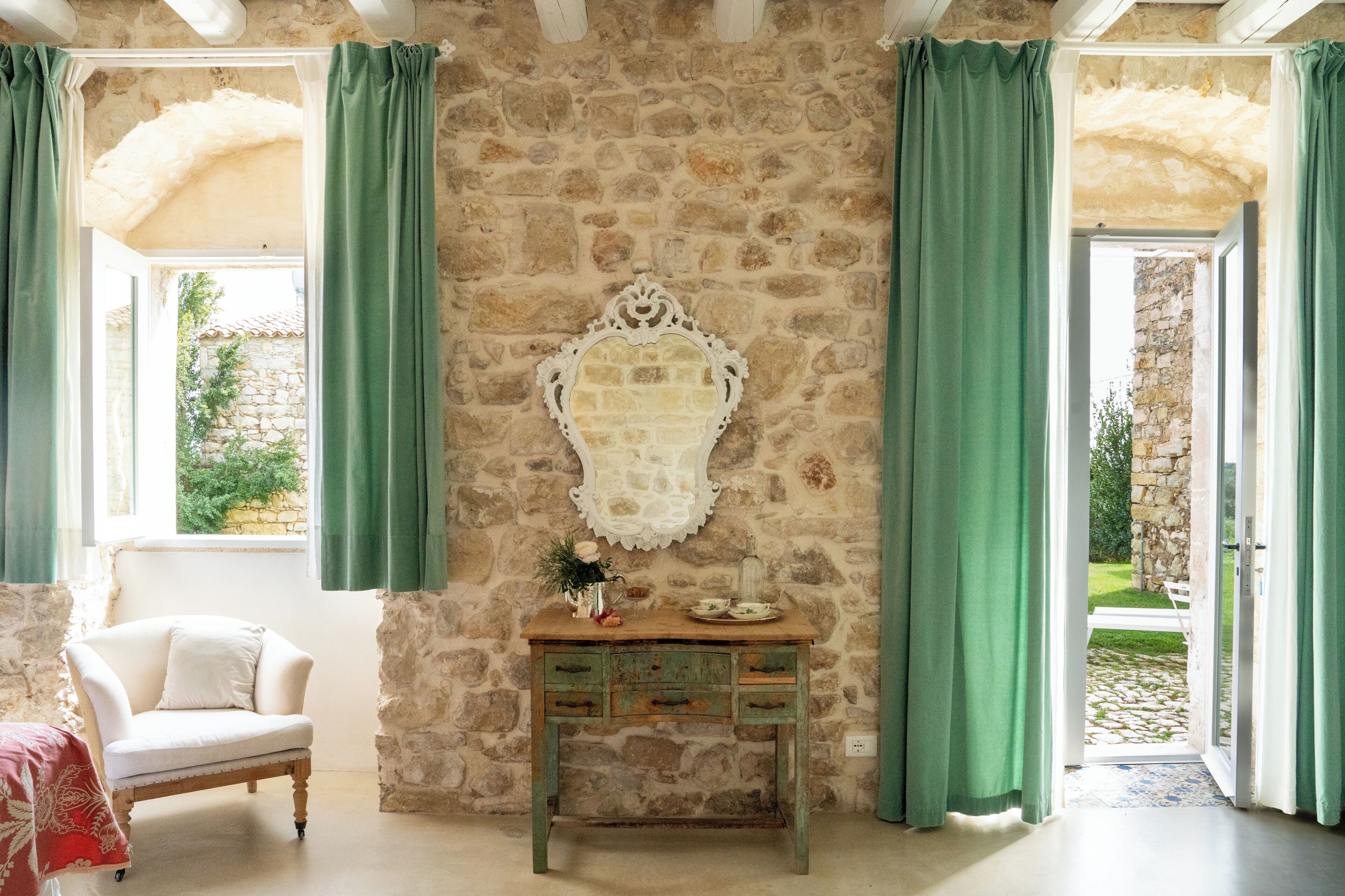 Una stanza con pareti in pietra, una sedia bianca, un tavolino consolle in legno con cassetti e uno specchio decorativo bianco.