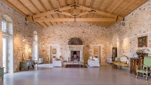 Amplia habitación con un alto techo de madera, paredes de piedra y una chimenea. Hay dos sofás blancos, un aparador de madera y un sillón verde.