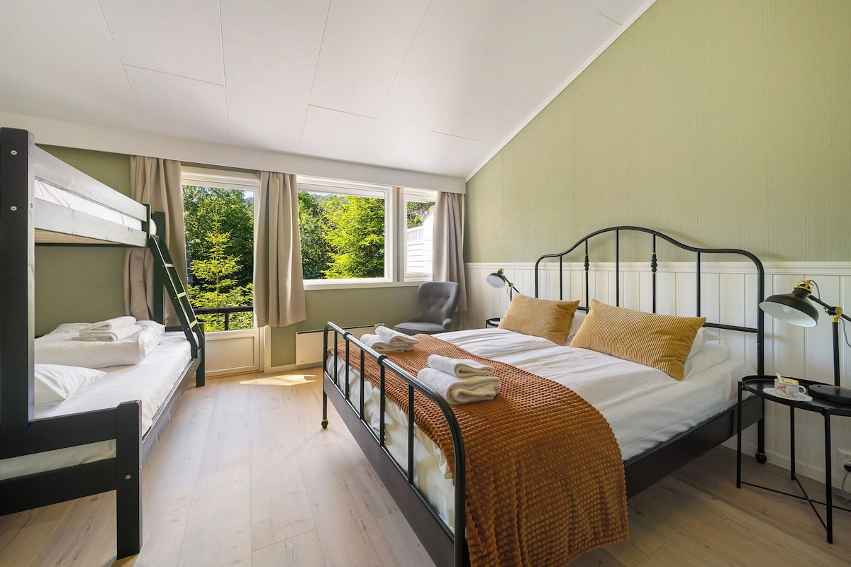 A bedroom with bunk beds, a single bed, a chair, a table with a lamp, and a view of greenery outside.