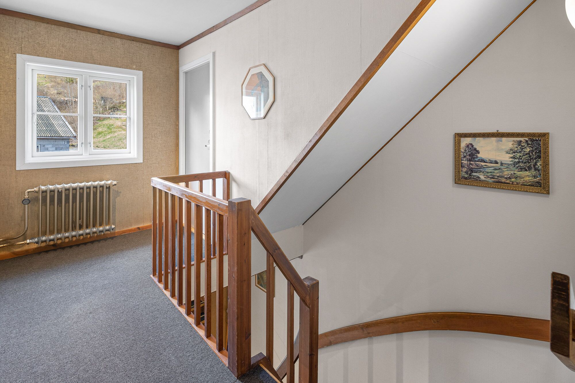 Un escalier avec des rampes en bois mène à un palier. On aperçoit un radiateur sur le sol, et une fenêtre donnant sur une maison se trouve à gauche.