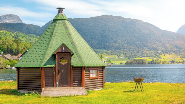 Una cabaña de troncos con techo verde y puerta de madera, ubicada junto a un lago con montañas al fondo.