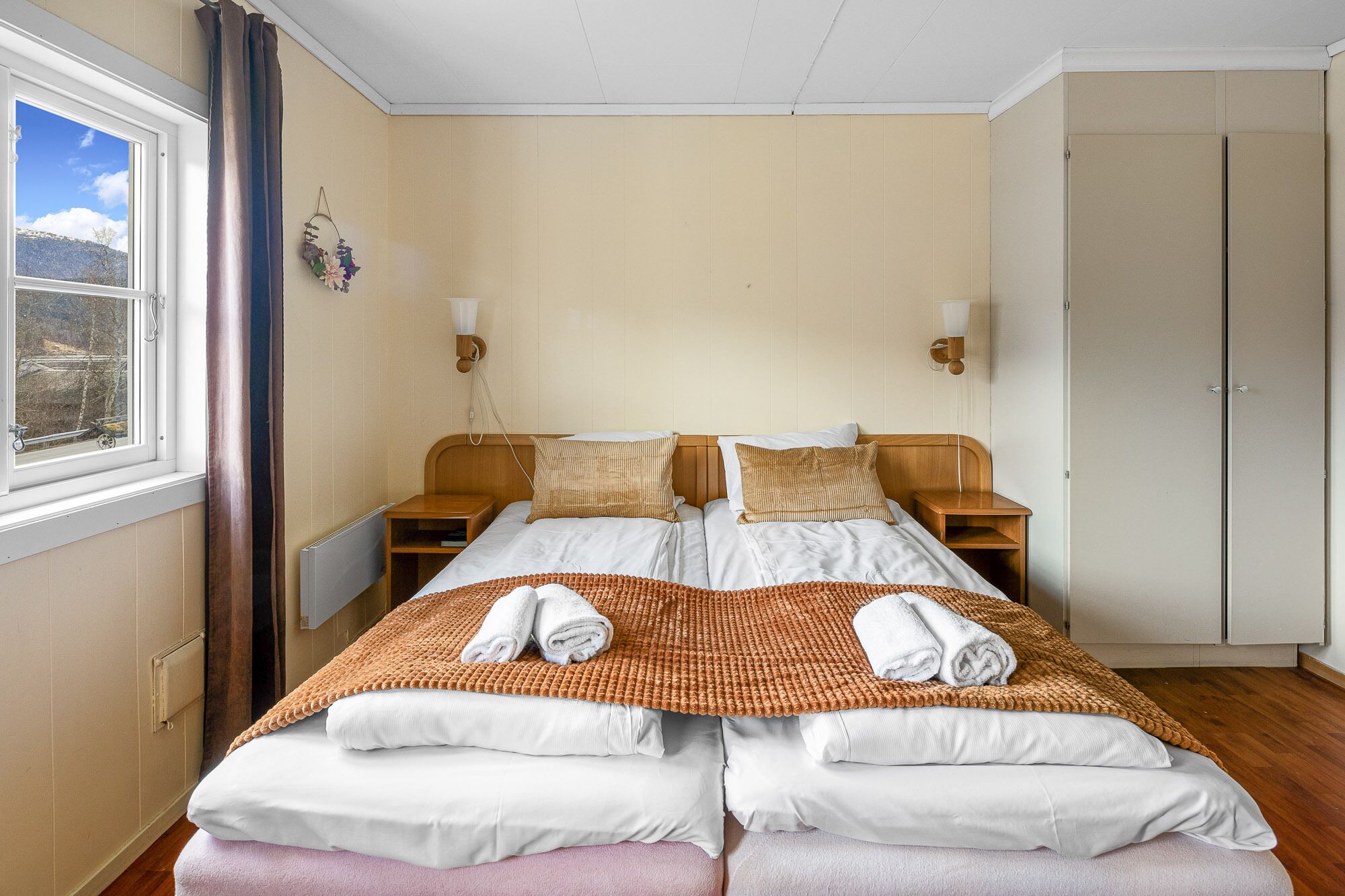 A double bed with white and brown bedding, two bedside tables, a wall-mounted lamp, and a view of the outdoors through a window.