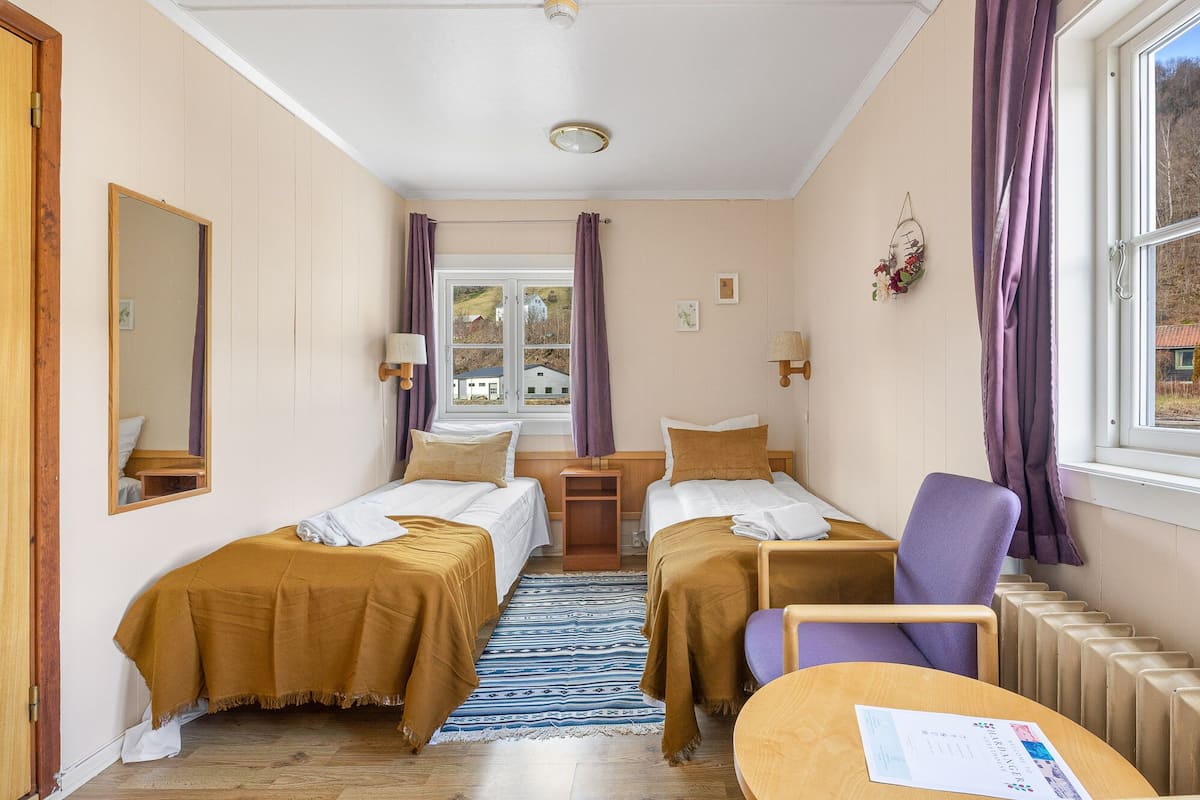 A hotel room with two beds, a window with curtains, a mirror, a small table, and a radiator.