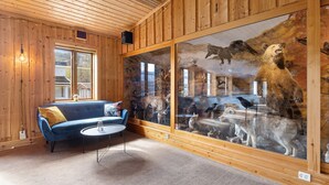 Una habitación con paredes de madera, un sofá azul, una mesa redonda y un amplio ventanal que muestra animales disecados.