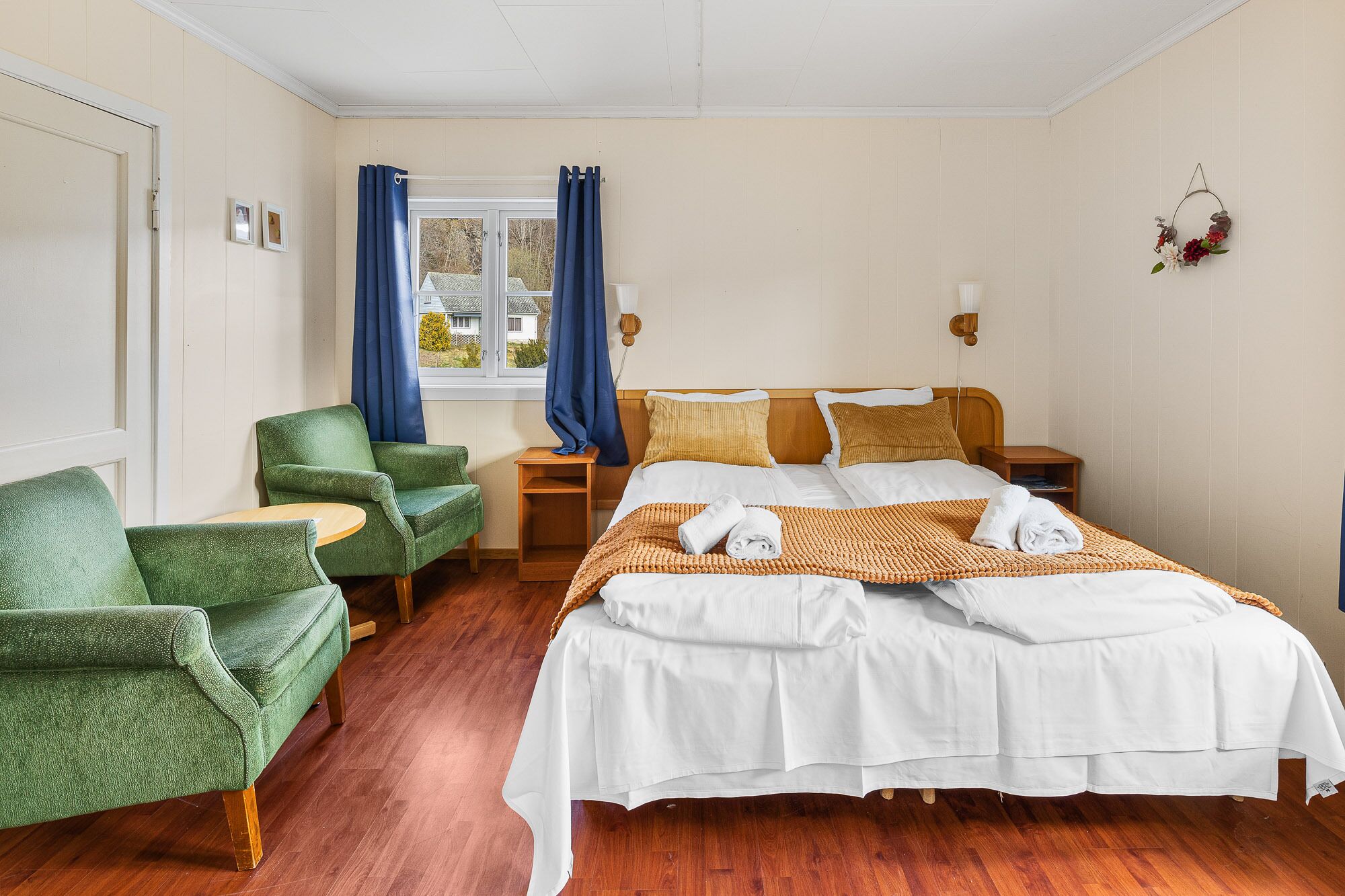 Une chambre d’hôtel avec un lit, un fauteuil vert, une table de chevet en bois et une fenêtre avec des rideaux.