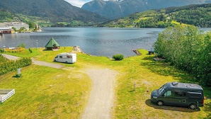 Un camping junto al lago con una cabaña de techo verde, una caravana blanca y una autocaravana oscura estacionados en una zona cubierta de césped.