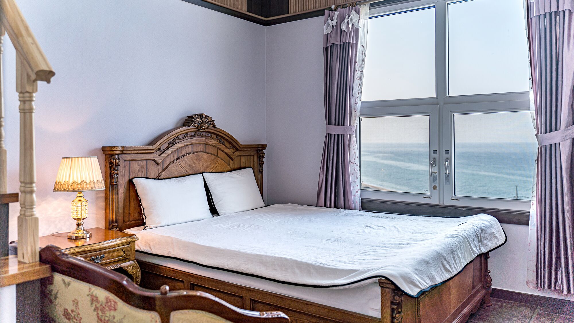 Un dormitorio con cama de madera, mesita de noche con lámpara y una ventana grande con cortinas.