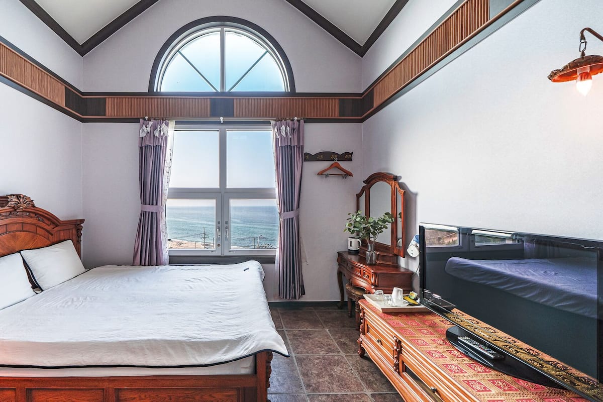 A bedroom with a large bed, a TV, a dresser, and a window with a view of the sea.