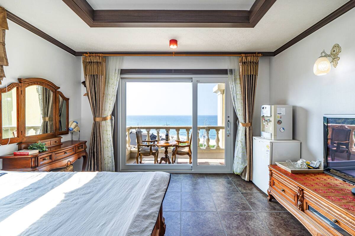 A hotel room with a bed, a television, a minibar, a water dispenser, and a view of the sea through the balcony doors.