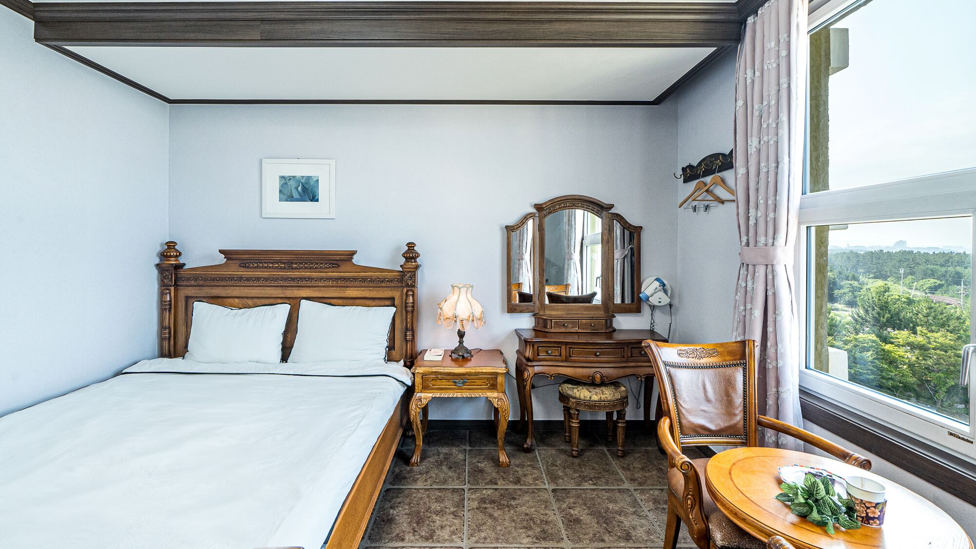Ein Schlafzimmer mit einem hölzernen Bett, einem Nachttisch, einer Kommode, einem Sessel und einem Tisch mit Tasse und Blättern.