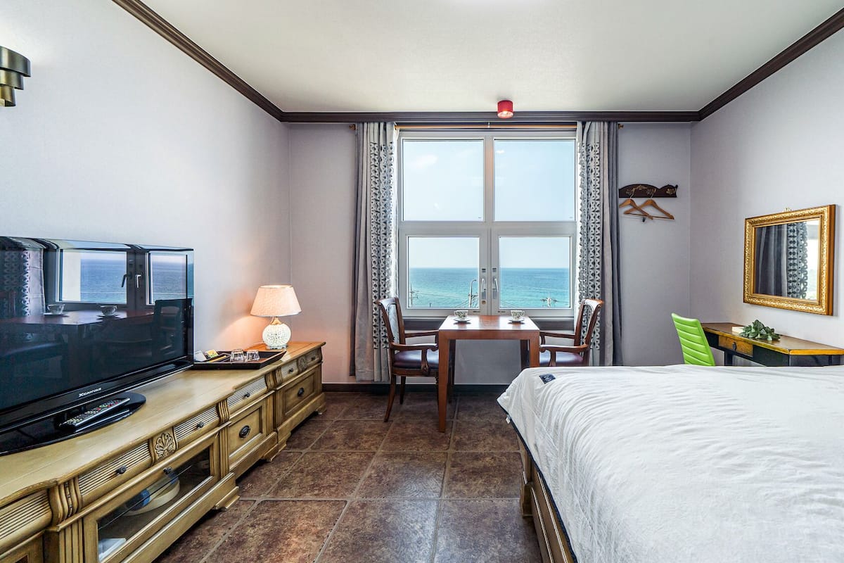 A hotel room with a bed, a TV, a desk with a chair, a table with two chairs, a window with a view of the sea, and a mirror on the wall.