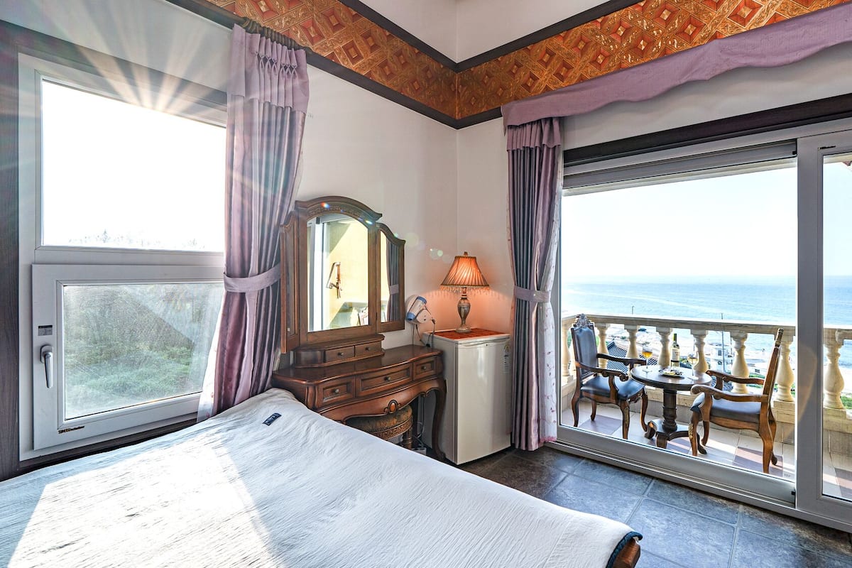A bedroom with a large bed, a dresser, a mirror, a lamp, and a balcony with a view of the sea.
