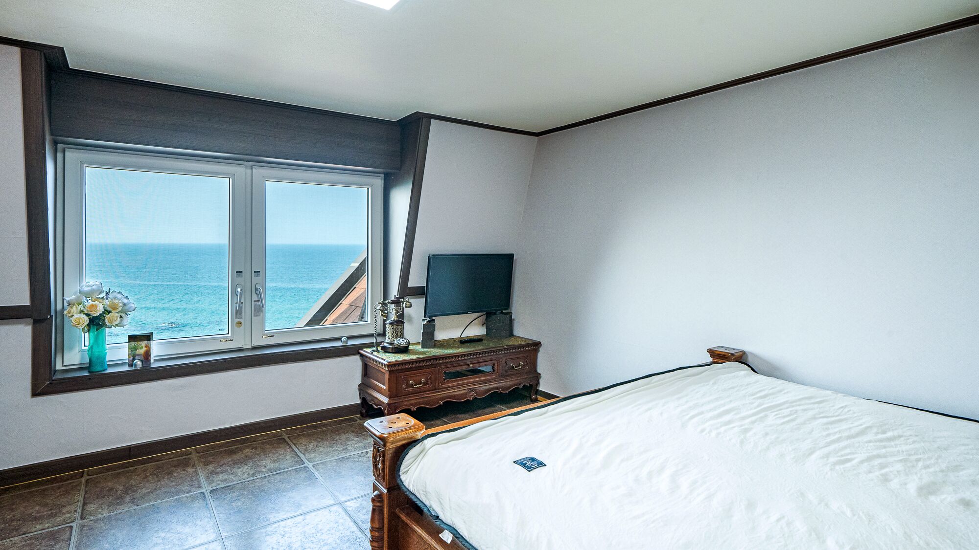 A bedroom with a bed, a nightstand, a television, and a window with a view of the sea.
