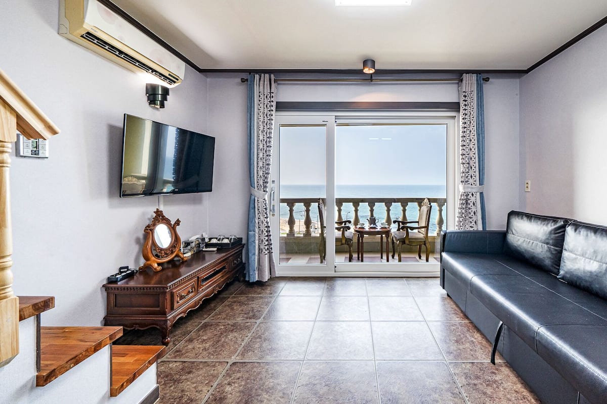 A living room with a black leather sofa, a TV mounted on the wall, a wooden cabinet, and a balcony with a view of the sea.