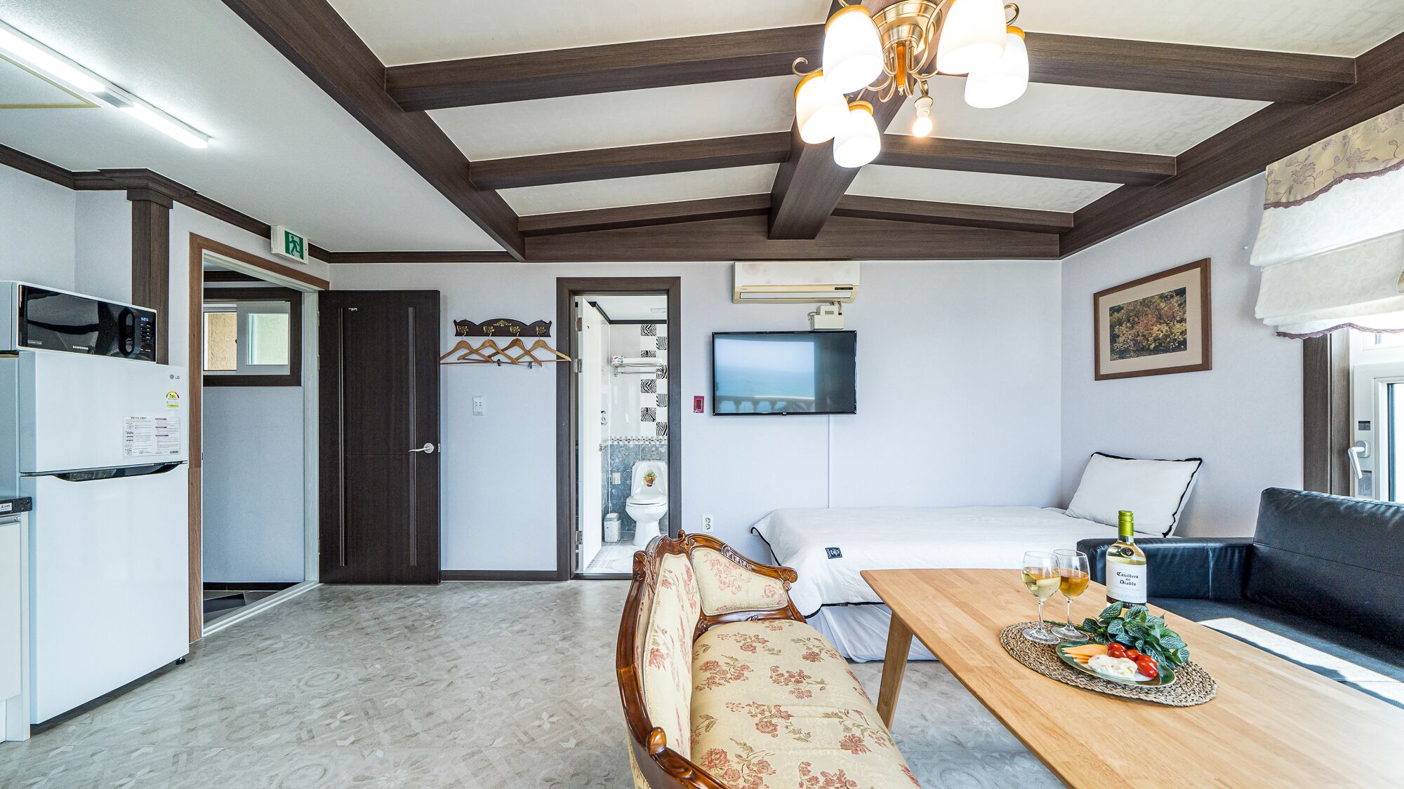 A modern hotel room with a wooden ceiling, a bed, a sofa, a dining table with a bottle of wine, and a TV on the wall.