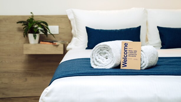 A neatly made bed with a rolled white towel and a welcome card, a potted plant, and a wooden nightstand with books. - Air Rooms Rome Airport by HelloSky (Fiumicino)