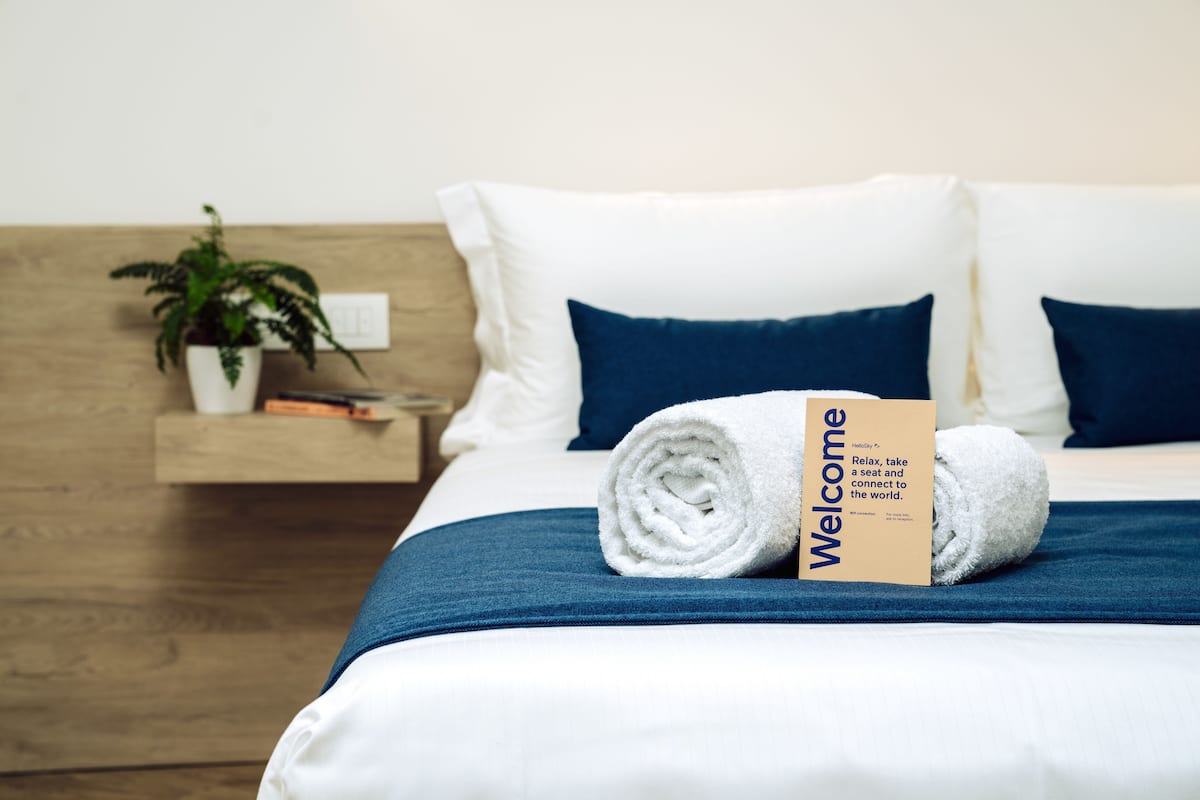 A neatly made bed with a rolled white towel and a welcome card, a potted plant, and a wooden nightstand with books.