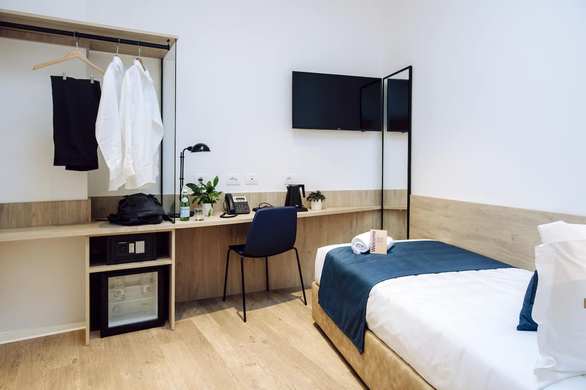 A hotel room with a bed, a desk with a chair, a TV mounted on the wall, and a wardrobe with clothes hanging.
