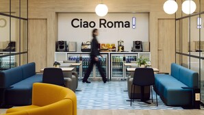 A modern lounge area with blue and yellow seating, a coffee station, and a wall with the text "Ciao Roma!". - Air Rooms Rome Airport by HelloSky (Fiumicino)