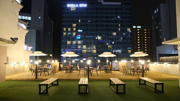 An outdoor seating area with umbrellas, benches, and tables, with a cityscape and a building labeled "SHILLA STAY" in the background. - Busan Haeundae Travel Pension (Busan)