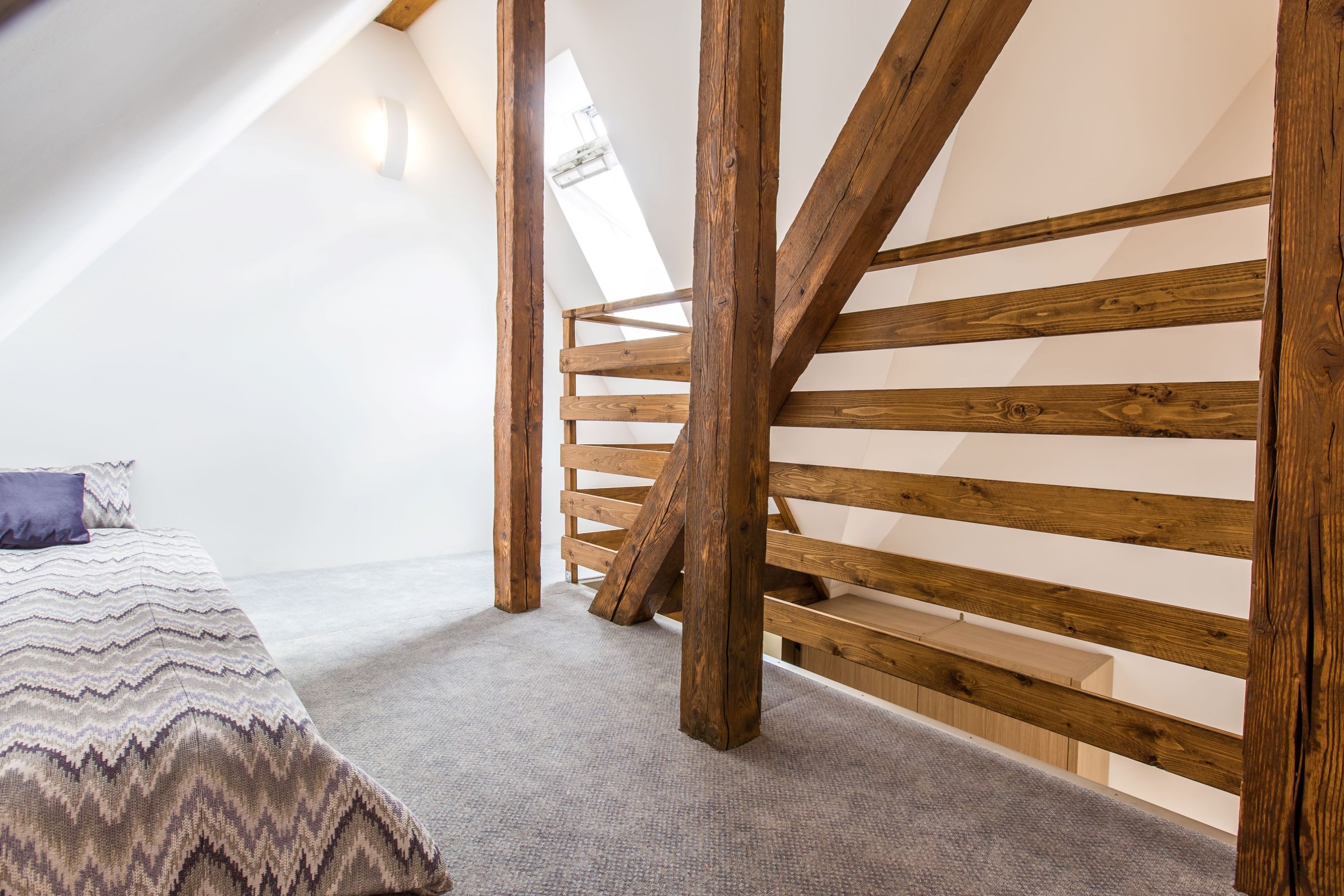 A bedroom with a sloped ceiling, wooden beams, a bed with patterned bedding, and a carpeted floor.