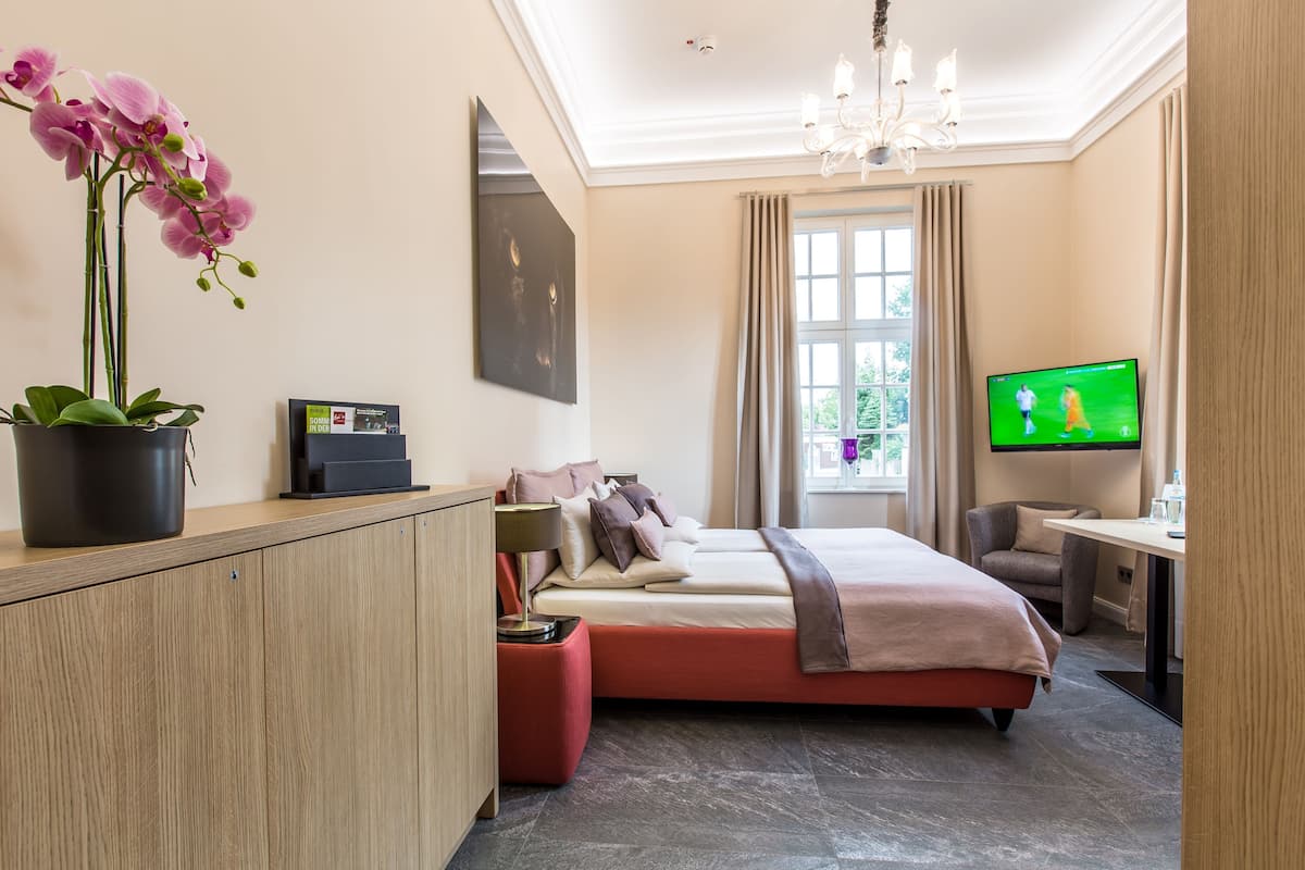 A hotel room with a bed, a desk, a TV, a chair, and a potted orchid.